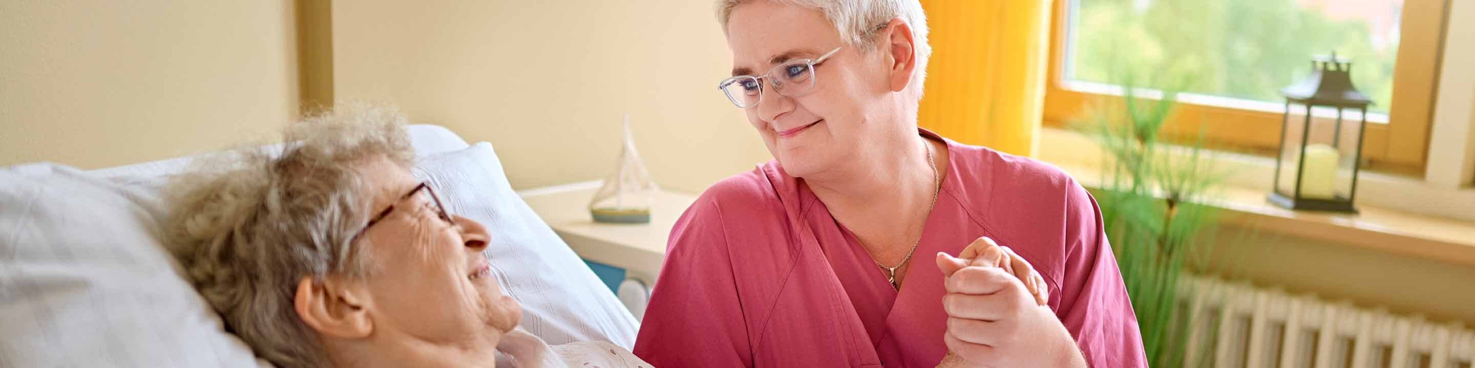Eine Pflegekraft hält die Hand einer älteren Patientin im Krankenhausbett und beide lächeln sich an.