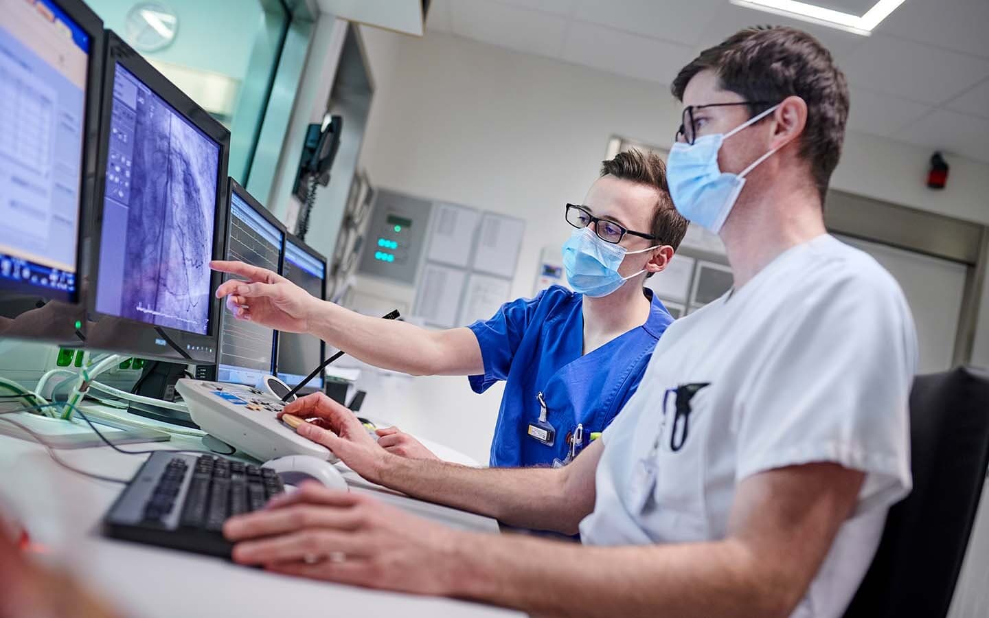 Zwei medizinische Fachkräfte mit Mundschutz betrachten mehrere Monitore und besprechen eine diagnostische Bildaufnahme aus dem Herzkatheterlabor.