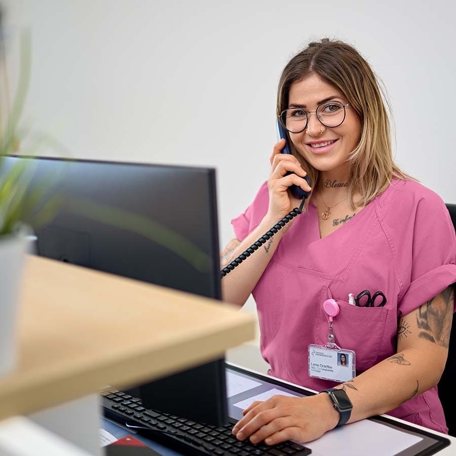 Mitarbeiterin des Krankenhauses sitzt vor einem PC, telefoniert und schaut dabei in Richtung der Kamera.