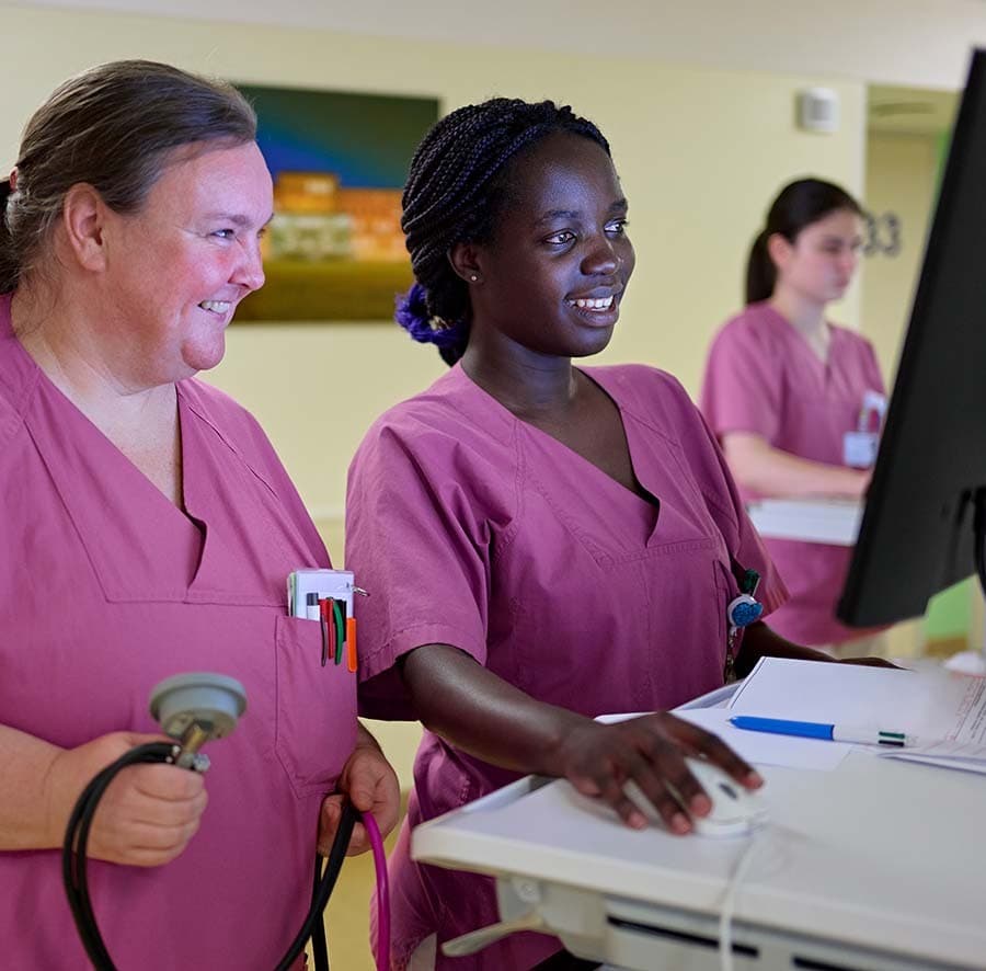 Eine Auszubildene der Pflege bedient auf einer Bettenstation einen PC und wird von einer Kollegin eingewiesen.